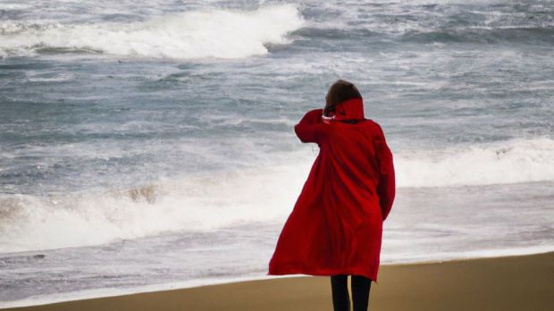 En una playa desierta, una mujer con un abrigo rojo se encuentra de pie frente al mar embravecido. El viento sopla con fuerza y las olas se estrellan contra los acantilados.